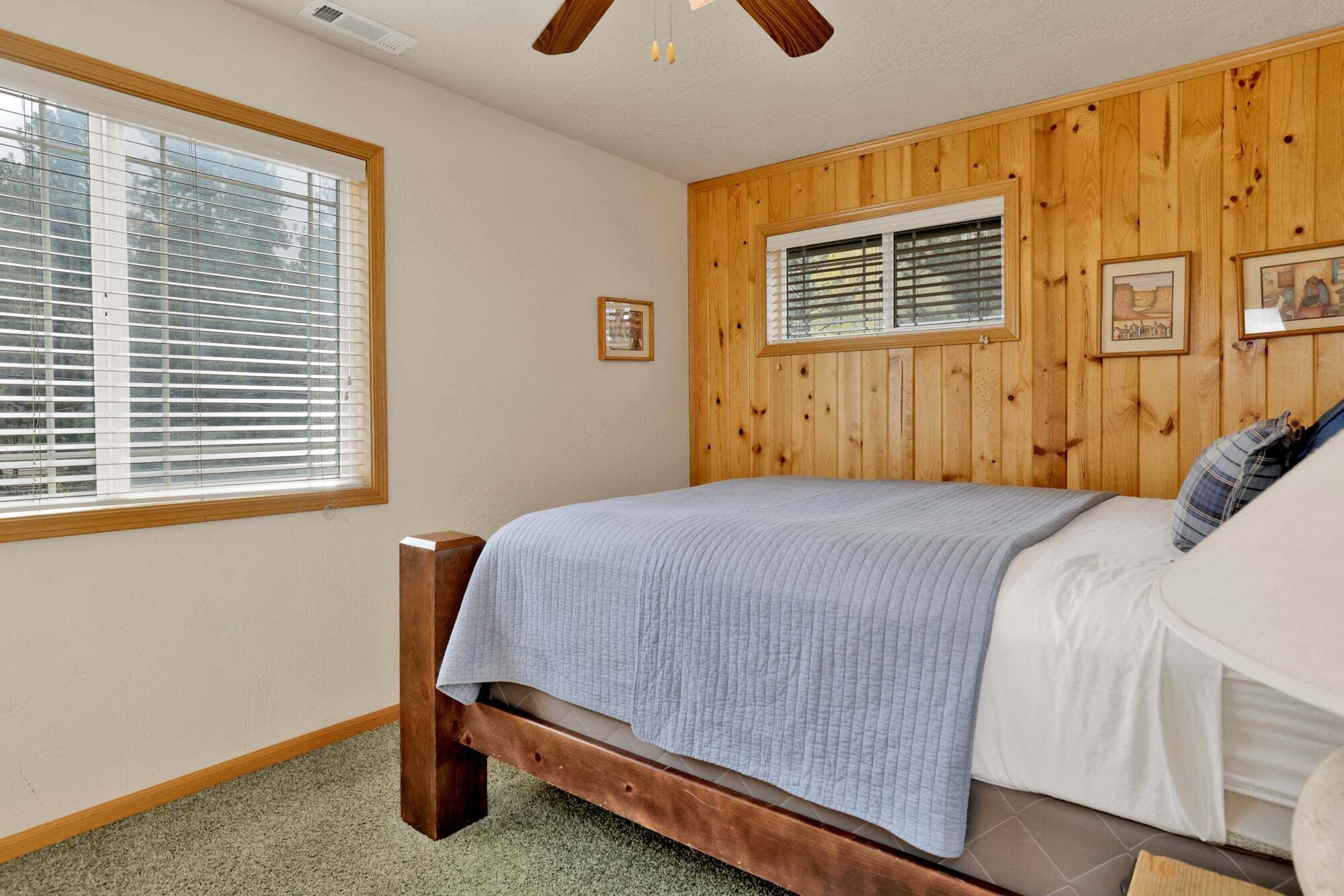 Un dormitorio con un revestimiento de madera, una cama con manta gris, una ventana con persianas y un ventilador de techo.