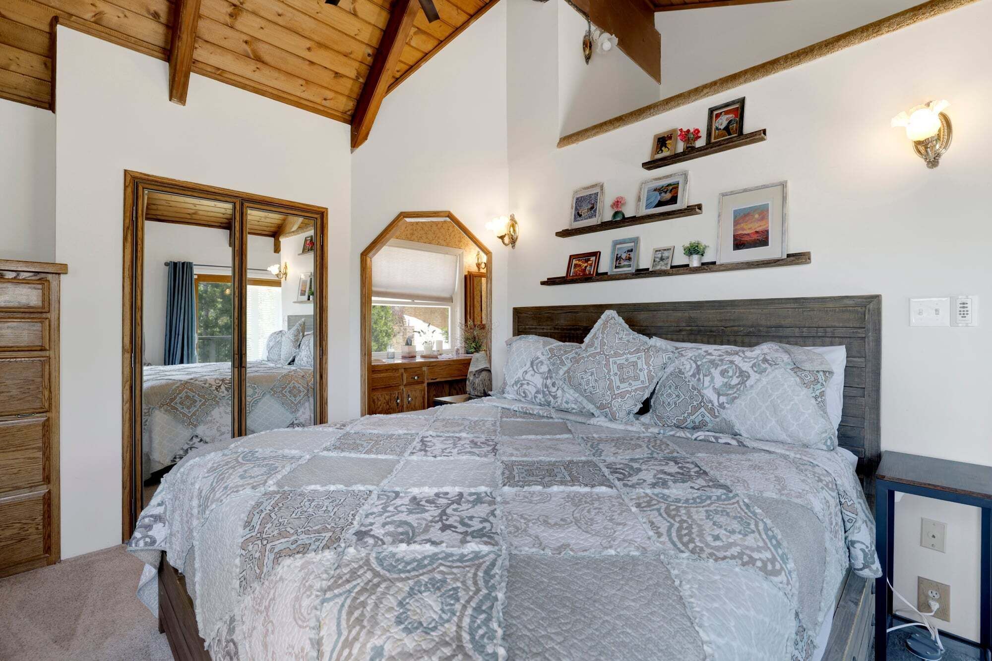 A bedroom with a large bed, wooden headboard, a mirror, and a shelf with pictures.