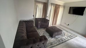 A living room with a sofa, ottoman, and armchair, a flat-screen TV, and a patterned rug. - Charming 3-bedroom House Rental in Centurion, South Africa (Centurion)