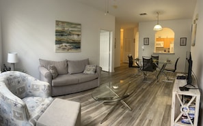 A living room with a grey sofa, a glass coffee table, and a patterned armchair. There is a dining area with a glass table and chairs, and a kitchen in the background. - Charming 2-bedroom apartment with AC in Kissimmee (Kissimmee)