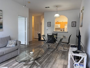 A modern living room with a glass coffee table, a gray sofa, and a dining area with a glass table and chairs. - Charming 2-bedroom apartment with AC in Kissimmee (Kissimmee)