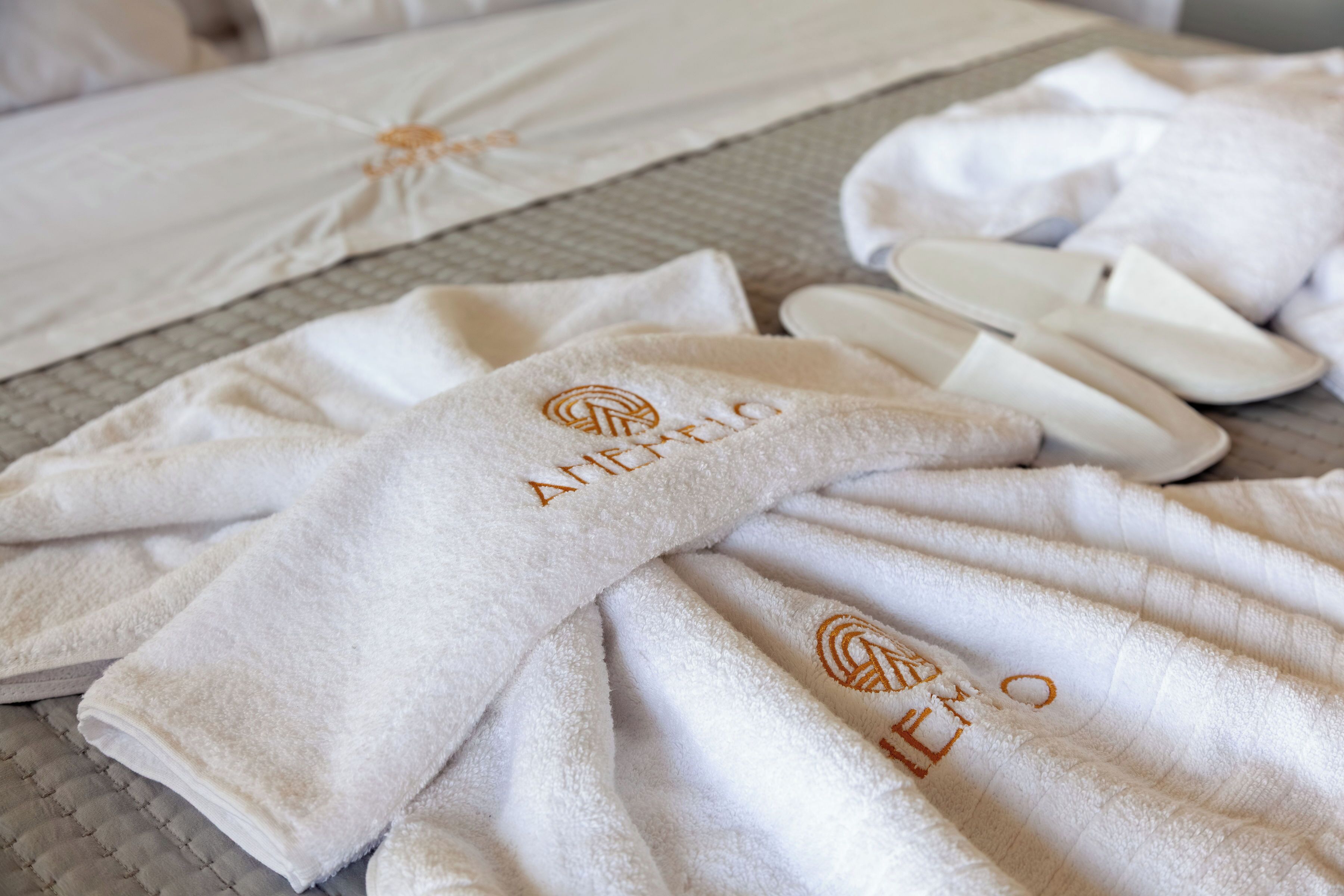 Toallas blancas bien dobladas con texto bordado, un par de pantuflas blancas y una cama con cubrecama acolchada.