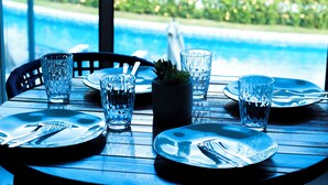 A dining table with plates, cutlery, and glasses, set against a view of a swimming pool. - FLAT BEACH CLASS SUMMER - PORTO (Ipojuca)