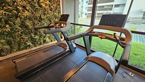 A treadmill with a digital display, a wall-mounted exercise bike, and a view of a building through large windows. - FLAT BEACH CLASS SUMMER - PORTO (Ipojuca)