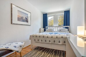 A neatly arranged bedroom with a bed, a bedside table, a lamp, a window with curtains, a framed picture on the wall, and a small bench.