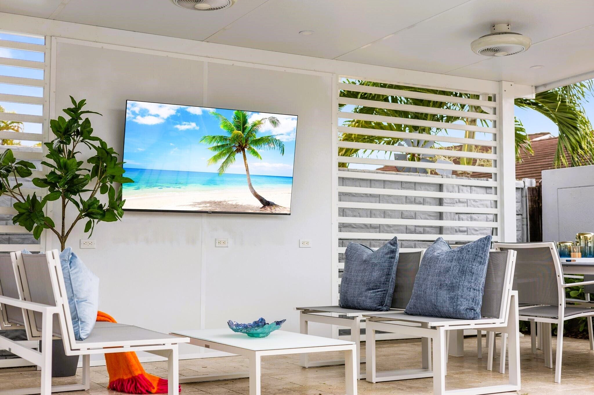 A modern outdoor seating area with white furniture, a large TV screen displaying a beach scene, and a potted plant.