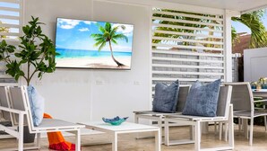Ett modernt utomhusområde med vita möbler, en stor TV-skärm som visar en strandscen och en krukväxt.