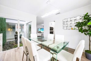 A modern dining area with a glass table and white chairs, a potted plant, and a wall-mounted decorative piece.