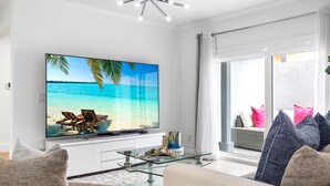 Ett modernt vardagsrum med en stor platt-TV som visar en strandscen, ett soffbord i glas och en soffa med kuddar.