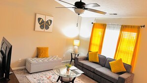 Ein Wohnzimmer mit Deckenventilator, einer gelben Kissensofa, einem Glastisch und einem gemusterten Teppich.