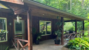 A wooden cottage with a covered porch, potted plants, and a view of the surrounding forest.