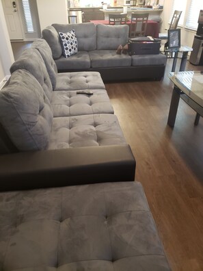 A sectional sofa with a remote control on it, a dining table, and a kitchen area in the background.