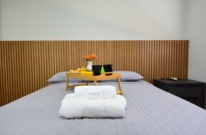 Un letto ben rifatto con coperta bianca, una testiera in legno, un vassoio con articoli da toilette e un tavolino con un vaso di fiori.