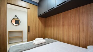 Cama bem arrumada com lençóis brancos, um cobertor cinza e duas toalhas enroladas. Há um guarda-roupa de madeira com espelho na parede e uma pequena estante de madeira.