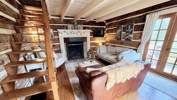 Interno di una baita di legno con camino, una scala, un divano e una porta che dà sull'esterno.