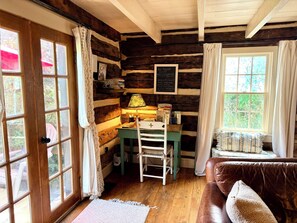 Ein rustikales Ferienhäuschen im Inneren, mit einer Holztür, einem Schreibtisch mit Lampe, einer Leiter, einer gepolsterten Bank und einem Fenster mit Vorhängen.