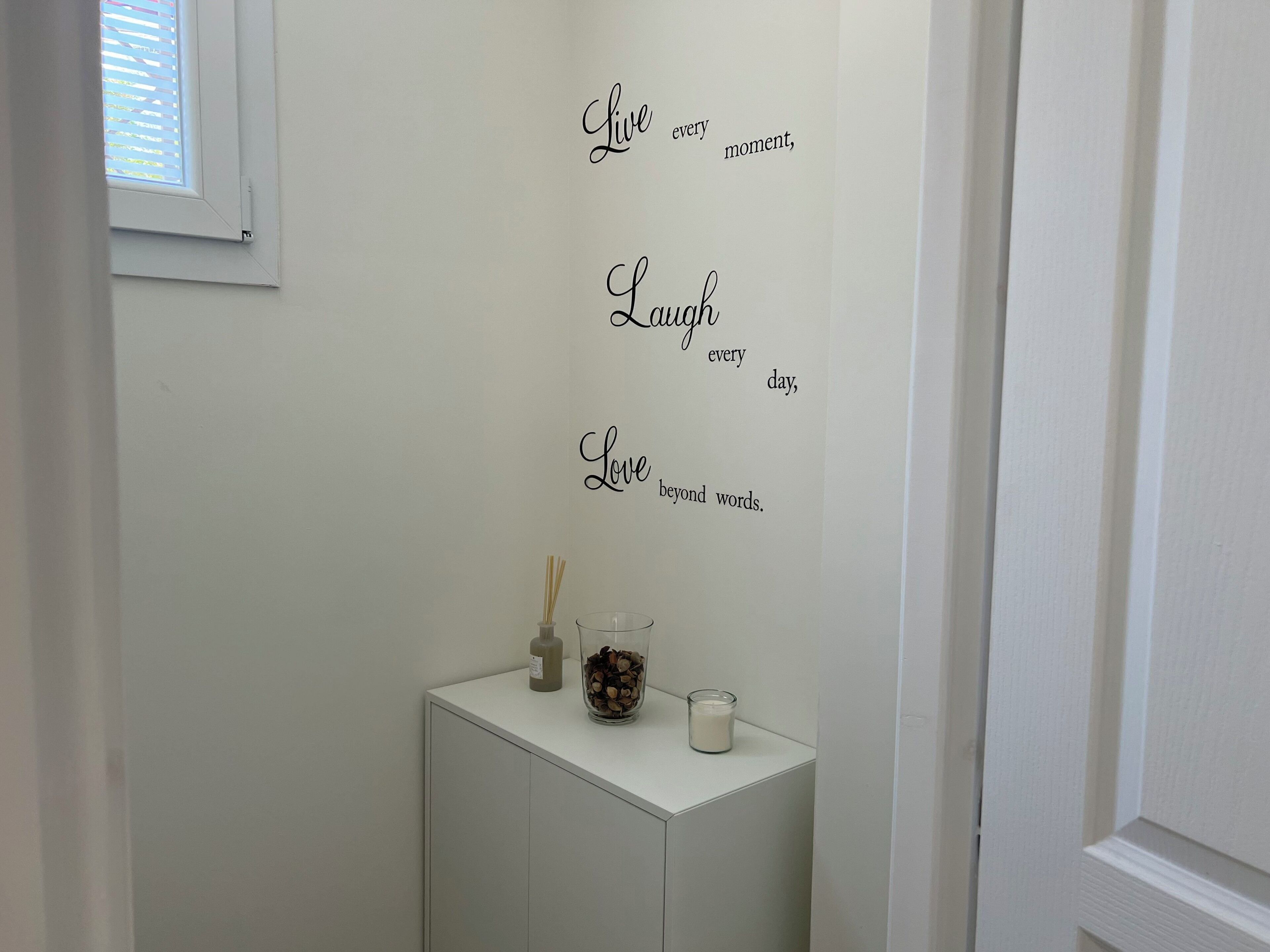 A hallway with a wall-mounted sign that reads 'Live every moment, Laugh every day, Love beyond words.' There is a small table with a bottle, a glass jar, and a candle.