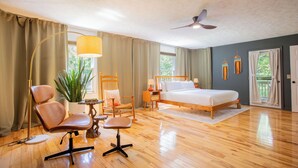 Ein Schlafzimmer mit einem Bett, zwei Stühlen, einem Schreibtisch, einer Lampe, einem Deckenventilator und einem großen Fenster.