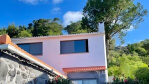 A two-story house with a stone facade, a tiled roof, and a small garden with pink flowers. - Casa da Petra – Our Casa is your Casa (Urzelina)