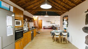 A kitchen with wooden cabinets, a refrigerator, and a microwave. There is a dining area with a table and chairs. - Casa da Petra – Our Casa is your Casa (Urzelina)