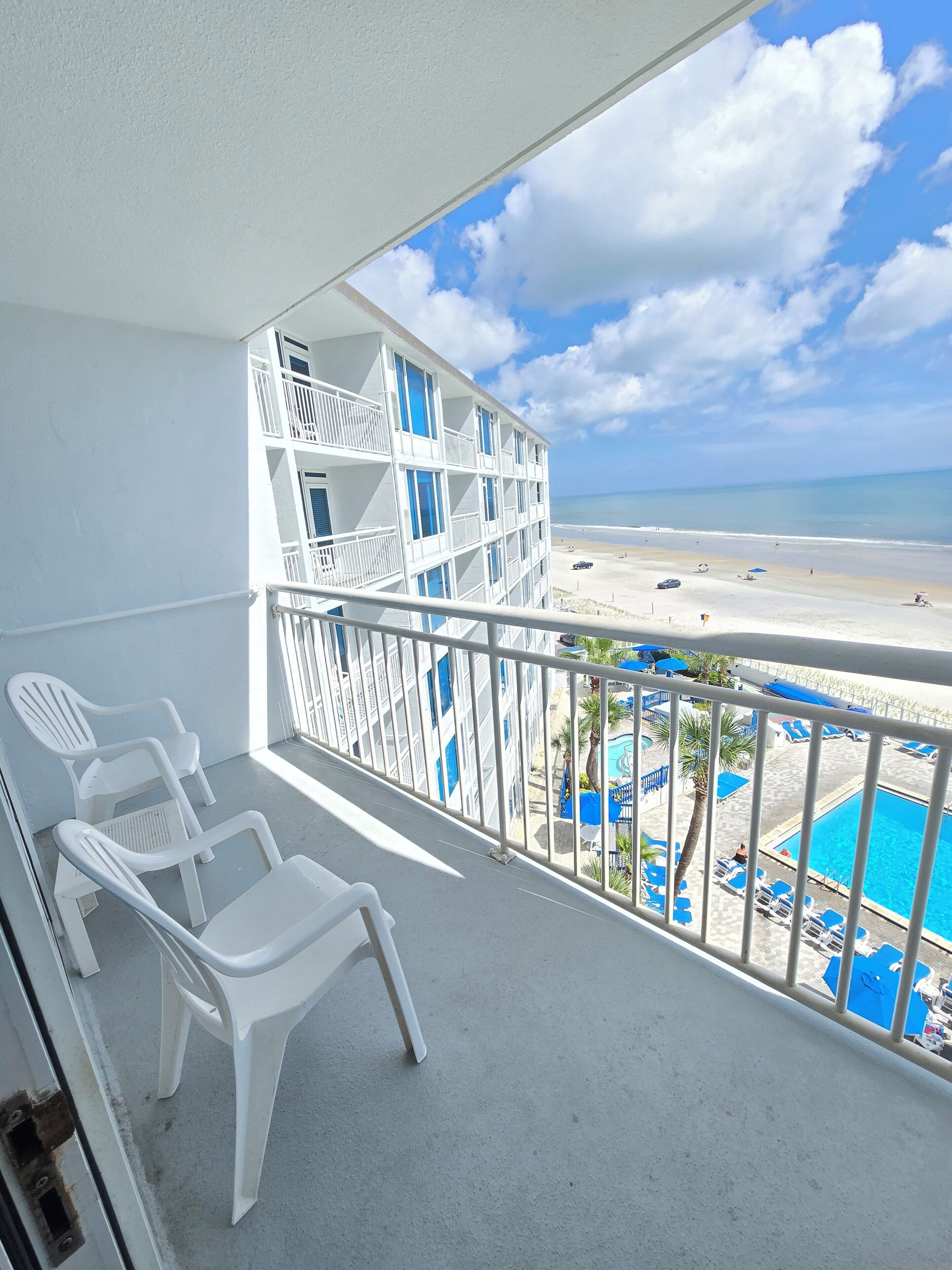 A balcony with white plastic chairs, a pool area with blue umbrellas, and a view of the beach and ocean.