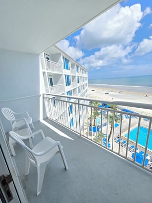 A balcony with white plastic chairs, a pool area with blue umbrellas, and a view of the beach and ocean. - Oceanfront Balcony - Pool - Islander Beach Resort, New Smyrna Beach! (New Smyrna Beach)
