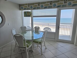 A dining area with a glass table and white wicker chairs, overlooking a beach.