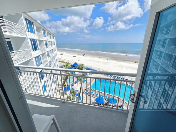 A balcony with a view of a beach, a pool, and a building with multiple floors.