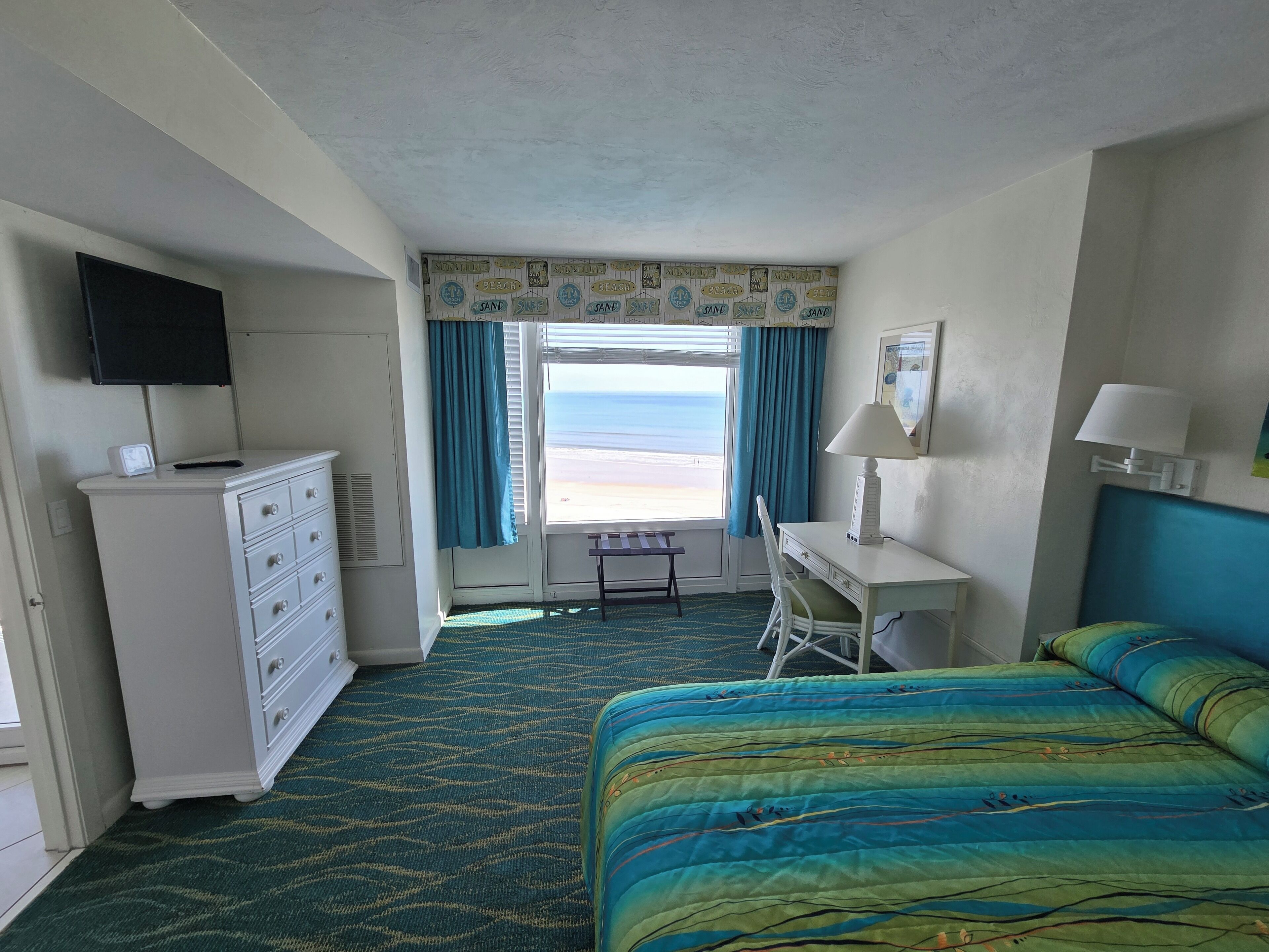 Habitación de hotel con cama, escritorio, cómoda, televisor y vista a la playa.