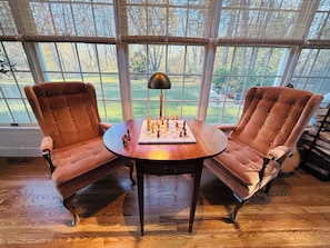 A room with two plush armchairs, a round wooden table with a chessboard, and a large window with a view of a green landscape.