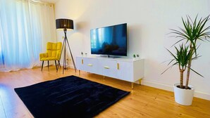Una sala de estar con alfombra negra, un sillón amarillo, un televisor sobre un mueble blanco, una lámpara de pie y una planta en maceta.