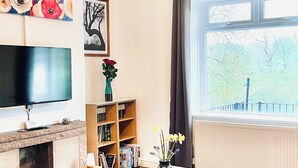 A living room with a flat-screen TV, a fireplace, a bookshelf, a table with flowers, and a large window with a view of trees. - DALE VIEW, Youlgreave, bright and cozy cottage in the heart of the Peak District (Youlgreave)