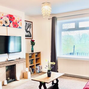 A living room with a flat-screen TV, a fireplace, a bookshelf, a table with flowers, and a large window with a view of trees. - DALE VIEW, Youlgreave, bright and cozy cottage in the heart of the Peak District (Youlgreave)