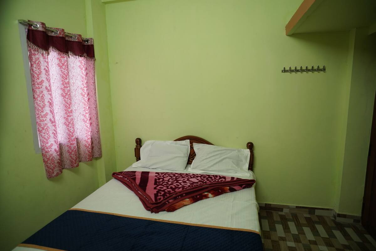 Una cama individual con cabecera de madera, manta estampada y ventana con cortina roja y blanca.