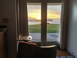A room with a view of the sunset over water, a door, a small table with a chair, and a rug on the floor. - Ty Mor (Newport)