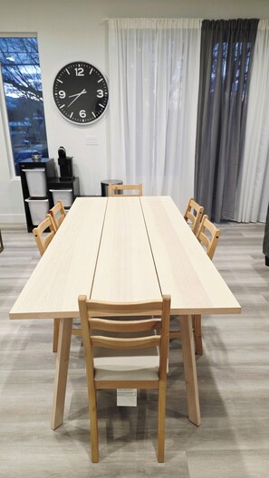 Un comedor con una larga mesa, seis sillas de madera, un reloj de pared, una ventana con vistas y una cortina.