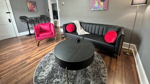 Een woonkamer met een zwarte leren bank, een rode fauteuil, een ronde salontafel en een schilderij aan de muur.