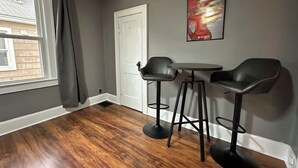 Een kamer met een houten vloer, een witte deur, twee zwarte stoelen, een klein rond tafeltje en een poster aan de muur.