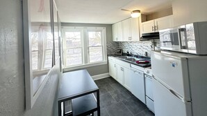 Een kleine keuken met witte kastjes, een roestvrijstalen koelkast en een raam met uitzicht op een besneeerd landschap.