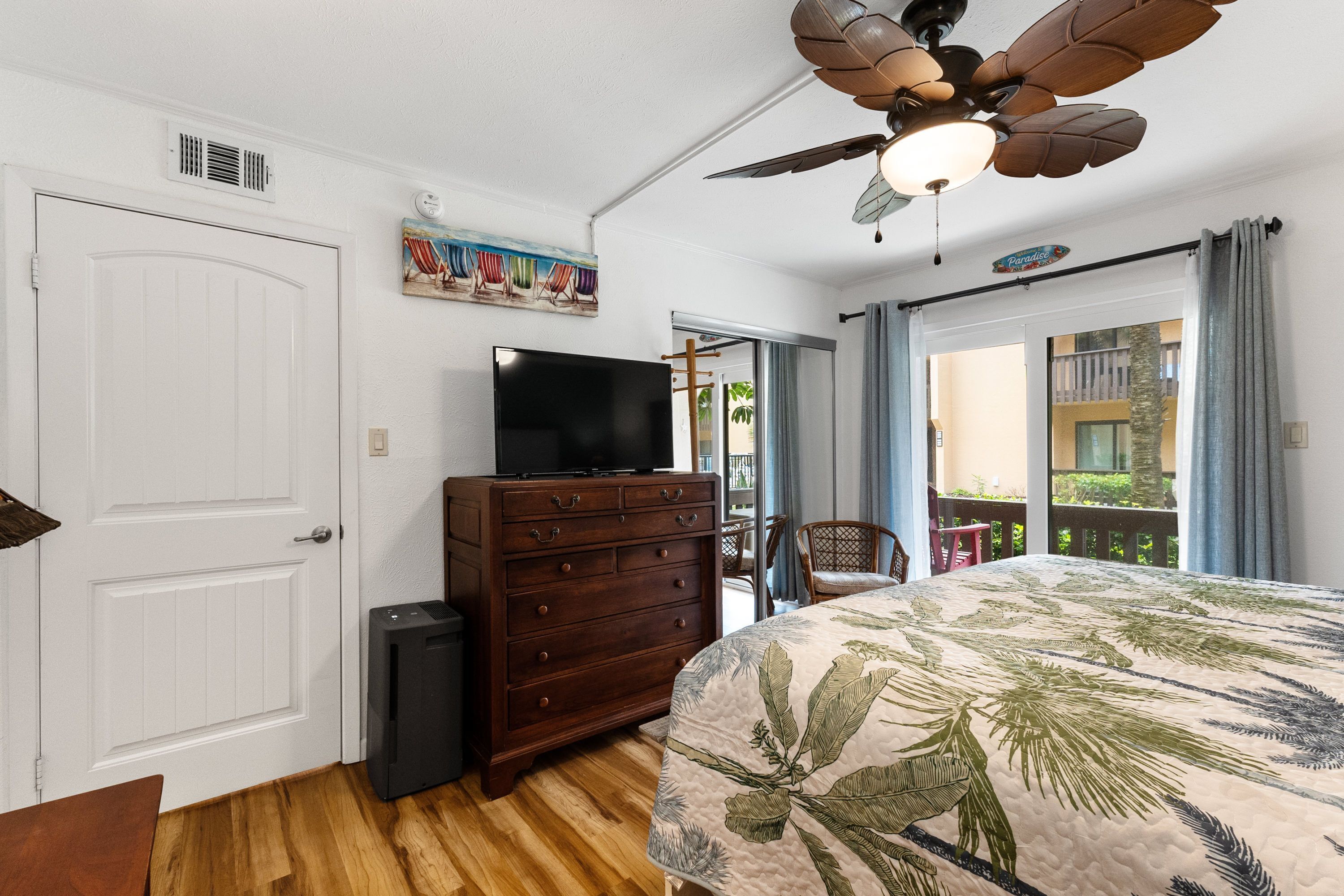 Une chambre avec un lit, une commode, un téléviseur, un ventilateur de plafond et une vue sur le balcon extérieur.