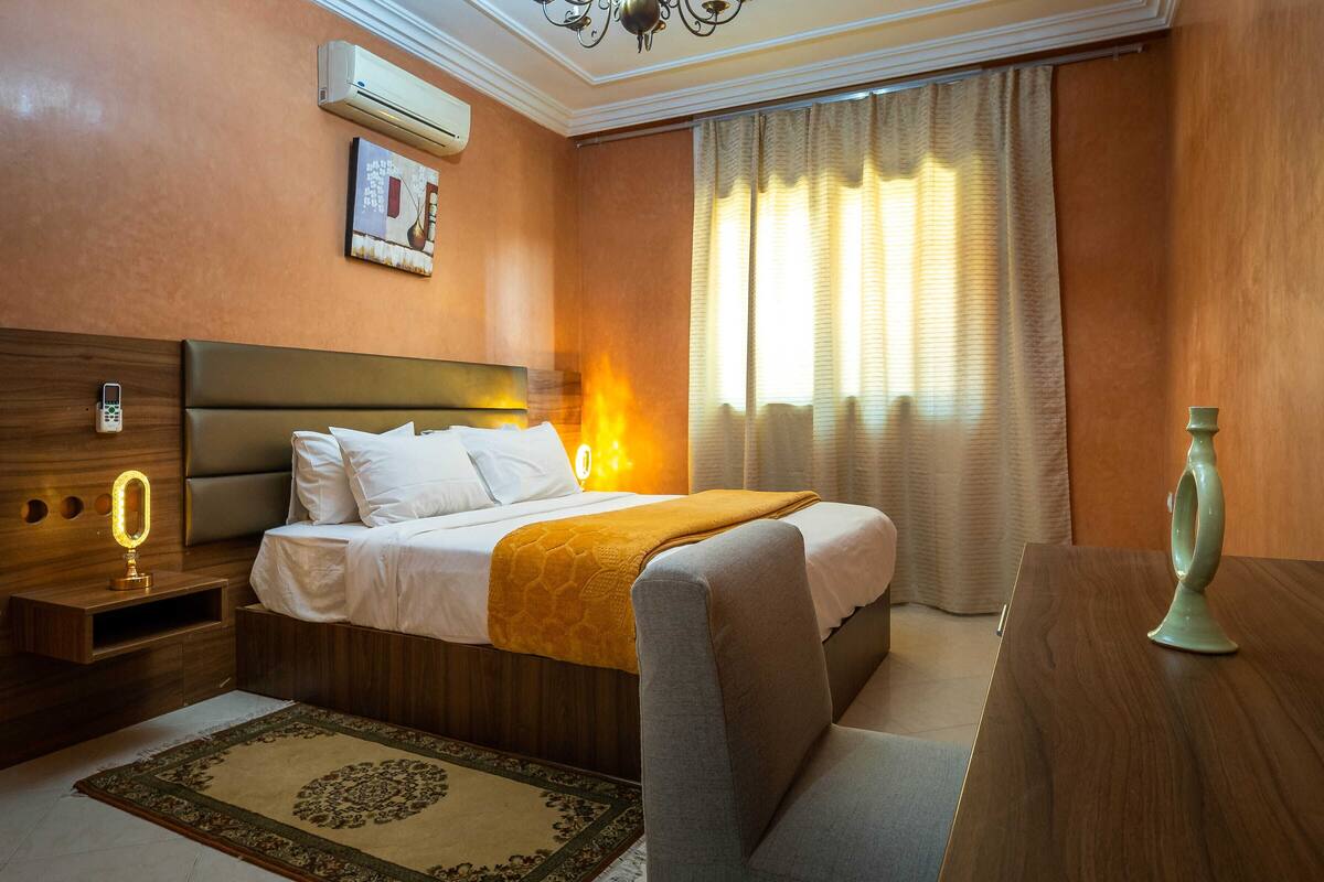 A hotel room with a bed, bedside table, lamp, and a large window with curtains.