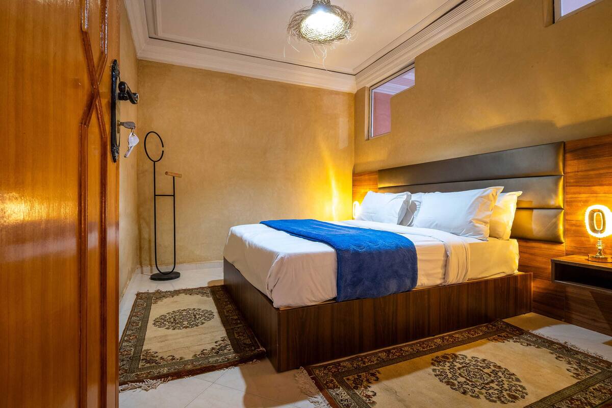A neatly made bed with a blue blanket, a bedside lamp, and a floor lamp in a room with beige walls and patterned rugs.