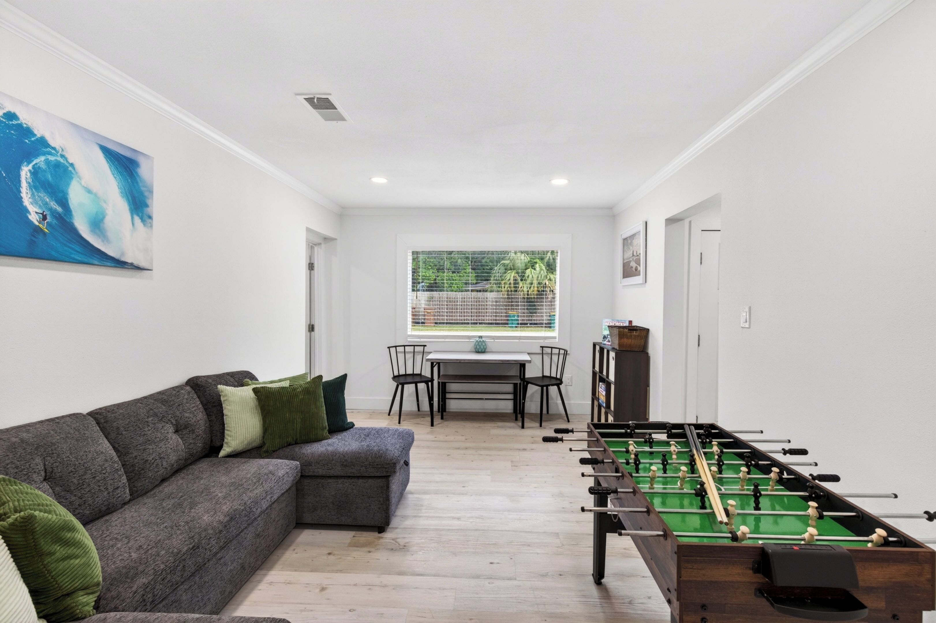 Een woonkamer met een bank, een tafelvoetbaltafel, een eettafel met stoelen en een raam met uitzicht op groen.