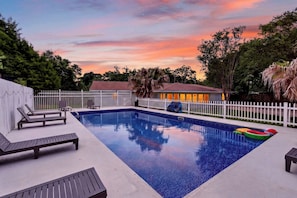 Una piscina rectangular con valla blanca, sillas de descanso y una casa al fondo.