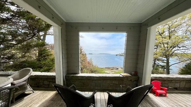 A covered porch with wicker chairs, a red chair, and a view of a lake. - Classic Maine Summer Cottage on the Ocean (Boothbay Harbor)
