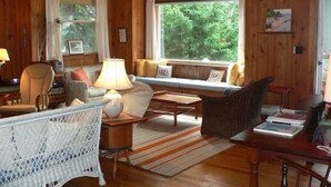 A cozy cabin interior with a sofa, chairs, a table, and a window with a view of greenery. - Classic Maine Summer Cottage on the Ocean (Boothbay Harbor)