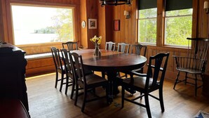 A wooden-paneled room with a round table and chairs, large windows, and a chandelier. - Classic Maine Summer Cottage on the Ocean (Boothbay Harbor)