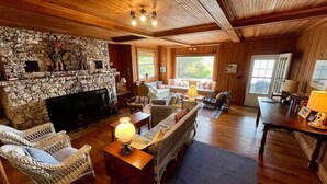 A cozy living room with a stone fireplace, wicker furniture, and a large window offering a view of the outdoors. - Classic Maine Summer Cottage on the Ocean (Boothbay Harbor)
