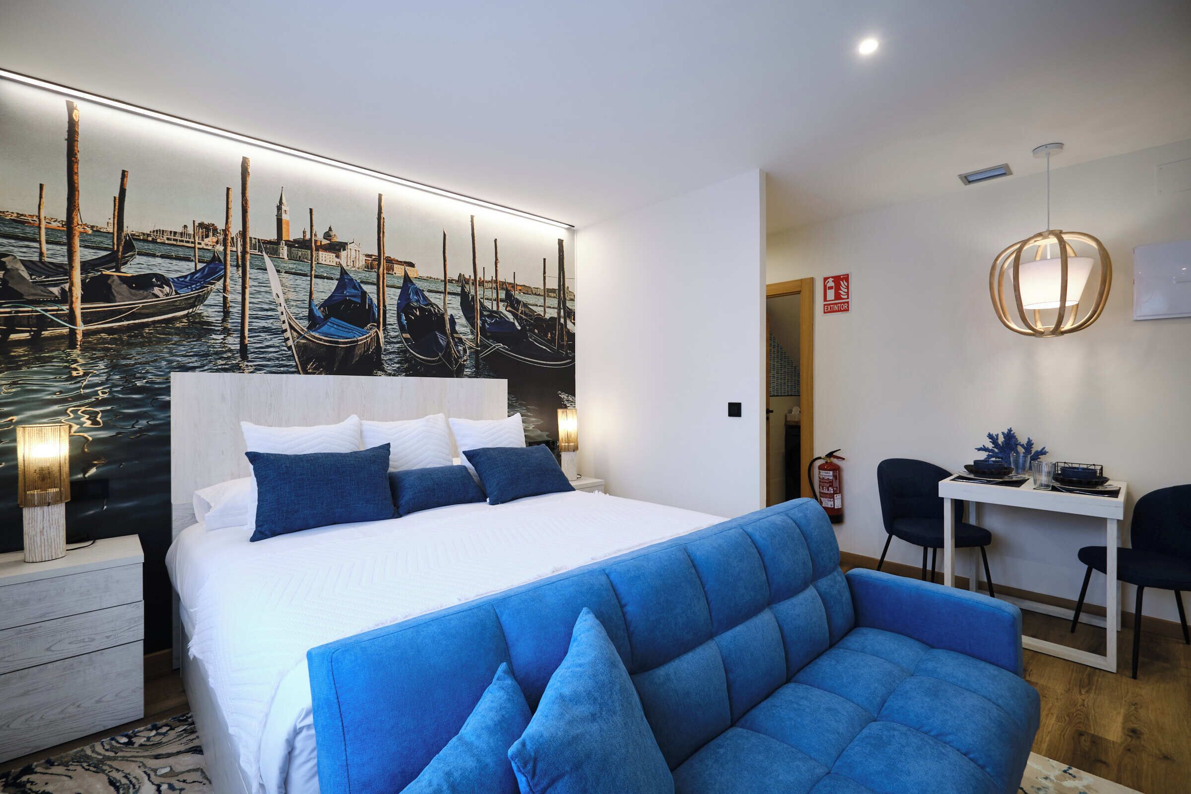 Una habitación de hotel con una cama, un sofá azul, un escritorio con dos sillas, una obra de arte en la pared con barcos y un extintor.