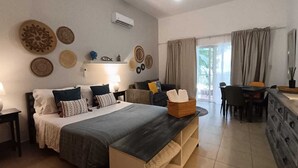 A bedroom with a bed, bedside tables, a dresser, a dining area, and a window with curtains. - Fastastico Apt. Al Cadaques villa and Resort (BAYAHIBE)